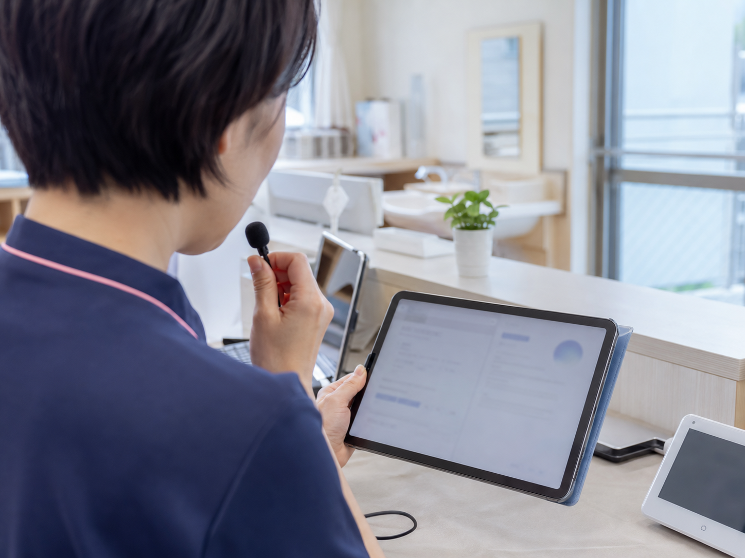 日本の介護施設で介護士がインカムマイクに音声入力しタブレットにAIの介護記録下書きが表示されている場面